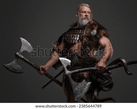 Mature gray-haired bearded Viking warrior in fur and light armor, wielding two enormous axes against a neutral background