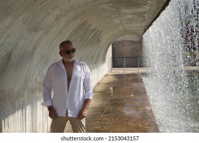 Mature, gray-haired man with beard, sunglasses, open white shirt and light brown pants, under a concrete tunnel next to a waterfall. Concept mature man, sugar daddy, lifestyle, teddy bear.