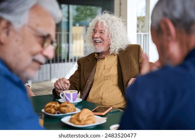Mature grey haired man smiling enjoying coffee with his group of friends on terrace of old people home. Meeting of senior Caucasian male persons sitting together having snack or breakfast outdoors