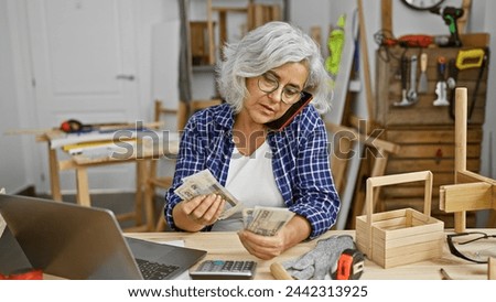 Mature grey-haired woman counting polish zloty bills in a carpentry workshop while talking on the phone.