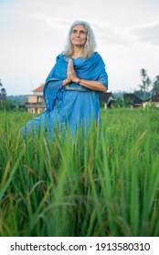 Mature grey-haired woman meditating standing in the yoga tree pose in a blue oriental dress nature indoors. Active healthy lifestyle sporty old person training workout wellness and outdoor exercising.