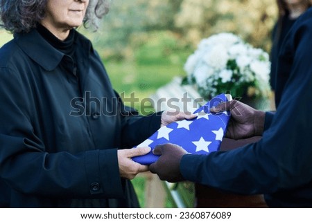 Mature grieving widow in black attire taking folded stars-and-stripes flag from hands of African American man at funeral of her husband