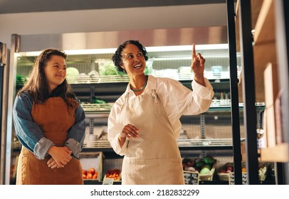 Mature Grocery Store Supervisor Giving Instructions To Her New Employee In A Shop. Female Small Business Owner Training A Woman With Down Syndrome As A Retail Sales Worker.