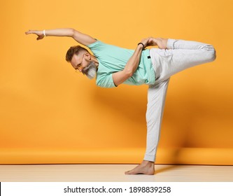 Mature Handsome Gray-haired Brutal Man In Fashionable Sportswear, Practicing Natarajasana, The Master Of Dance Yoga, Puts On Balance And Stretching. The Studio Is Filmed Against A Yellow Background