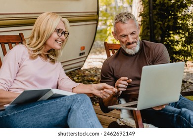 Mature handsome grey-haired man showing his blonde wife something amusing on his laptop in their garden, working remotely, freelancing online while traveling by trailer - Powered by Shutterstock