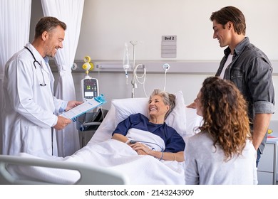 Mature Happy Doctor Giving Good News To Family And Old Patient. Smiling Senior Woman Lying On Hospital Bed With Son And Daughter Visiting And Talking To Physician. Doctor Visiting Hospitalized Patient