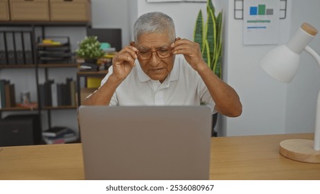 Mature hispanic man adjusting glasses while working on a laptop in a well-organized office space with shelves and plants in the background. - Powered by Shutterstock