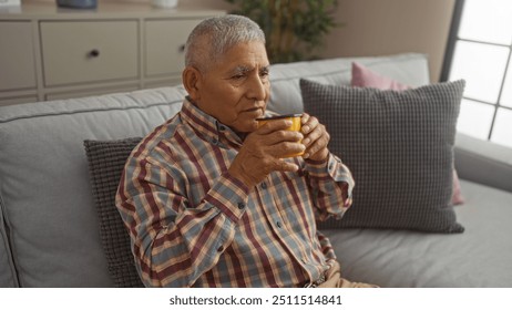 Mature hispanic man enjoying a hot drink indoors, sitting on a couch in a cozy living room with a plaid shirt and a thoughtful expression. - Powered by Shutterstock