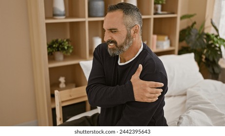 A mature hispanic man with grey beard in pain, holding his shoulder indoors, expressing discomfort in a bedroom setting. - Powered by Shutterstock