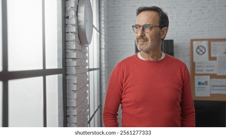 Mature hispanic man wearing glasses standing in an office with brick wall interior looking thoughtfully out the window in casual red sweater - Powered by Shutterstock