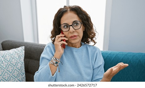 A mature hispanic woman with curly hair talking on a phone in a cozy living room, showing confusion or concern. - Powered by Shutterstock