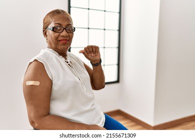 Mature Hispanic Woman Getting Vaccine Showing Arm With Band Aid Pointing To The Back Behind With Hand And Thumbs Up, Smiling Confident 