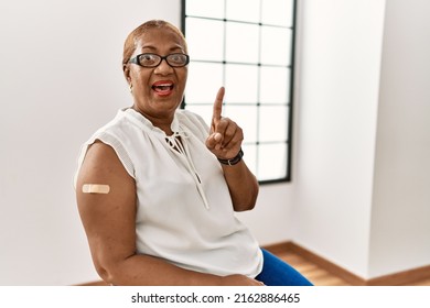 Mature Hispanic Woman Getting Vaccine Showing Arm With Band Aid Pointing Finger Up With Successful Idea. Exited And Happy. Number One. 