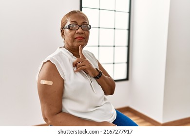 Mature Hispanic Woman Getting Vaccine Showing Arm With Band Aid Thinking Concentrated About Doubt With Finger On Chin And Looking Up Wondering 