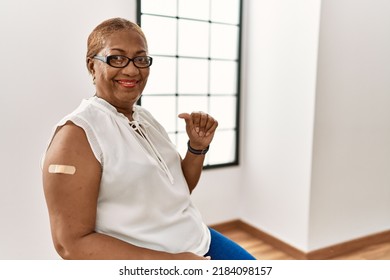 Mature Hispanic Woman Getting Vaccine Showing Arm With Band Aid Smiling With Happy Face Looking And Pointing To The Side With Thumb Up. 