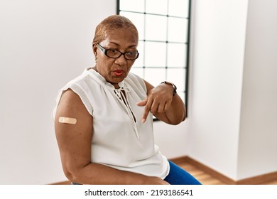 Mature Hispanic Woman Getting Vaccine Showing Arm With Band Aid Pointing Down With Fingers Showing Advertisement, Surprised Face And Open Mouth 