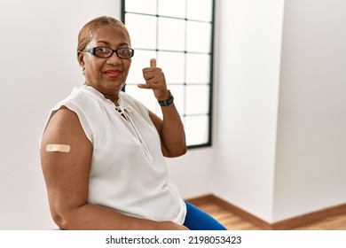 Mature Hispanic Woman Getting Vaccine Showing Arm With Band Aid Smiling Doing Phone Gesture With Hand And Fingers Like Talking On The Telephone. Communicating Concepts. 