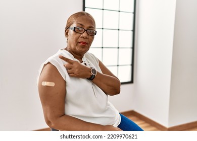 Mature Hispanic Woman Getting Vaccine Showing Arm With Band Aid Pointing With Hand Finger To The Side Showing Advertisement, Serious And Calm Face 