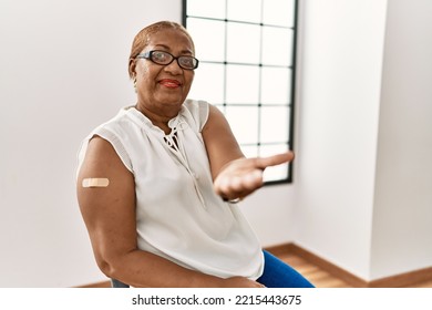 Mature Hispanic Woman Getting Vaccine Showing Arm With Band Aid Smiling Cheerful Offering Palm Hand Giving Assistance And Acceptance. 