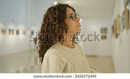Mature hispanic woman in glasses admiring art in a gallery interior, embodying elegance and contemplation.