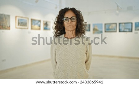 Mature hispanic woman with glasses standing thoughtfully in an art gallery, portraying a sense of contemplation and culture.