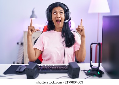 Mature Hispanic Woman Playing Video Games At Home Amazed And Surprised Looking Up And Pointing With Fingers And Raised Arms. 