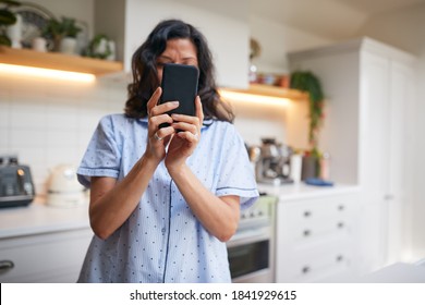 Mature Hispanic Woman In Pyjamas At Home In Kitchen Texting And Using Social Media On Mobile Phone