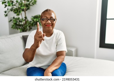 Mature Hispanic Woman Sitting On The Sofa At Home Showing And Pointing Up With Finger Number One While Smiling Confident And Happy. 