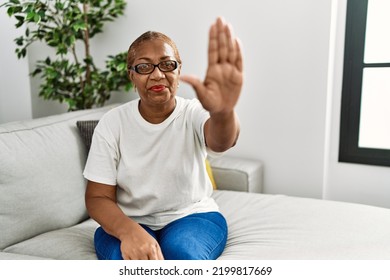 Mature Hispanic Woman Sitting On The Sofa At Home Doing Stop Sing With Palm Of The Hand. Warning Expression With Negative And Serious Gesture On The Face. 