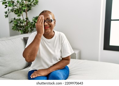 Mature Hispanic Woman Sitting On The Sofa At Home Covering One Eye With Hand, Confident Smile On Face And Surprise Emotion. 
