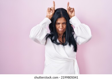 Mature Hispanic Woman Standing Over Pink Background Doing Funny Gesture With Finger Over Head As Bull Horns 