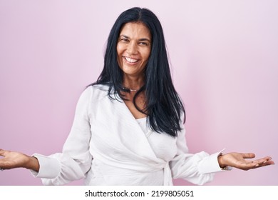 Mature Hispanic Woman Standing Over Pink Background Smiling Cheerful With Open Arms As Friendly Welcome, Positive And Confident Greetings 