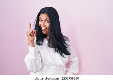 Mature Hispanic Woman Standing Over Pink Background Pointing Finger Up With Successful Idea. Exited And Happy. Number One. 