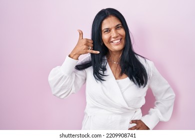 Mature Hispanic Woman Standing Over Pink Background Smiling Doing Phone Gesture With Hand And Fingers Like Talking On The Telephone. Communicating Concepts. 