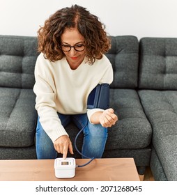 Mature Hispanic Woman Using Blood Pressure Monitor Sitting On The Sofa At Home