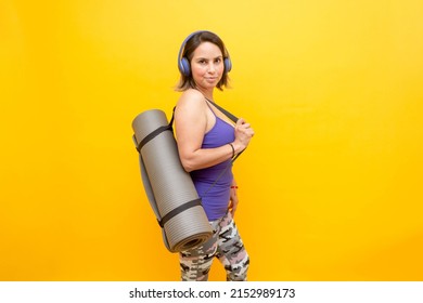 Mature Hispanic Woman Wearing Sportswear And Headphones, Standing Holding Yoga Mat