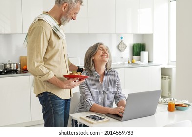 Mature Husband Bringing Breakfast To Happy Wife Working From Home Office. Older Mid Age Man Serving Meal Smiling Senior Woman Helping With Household While Spouse Using Laptop Sitting At Kitchen Table.