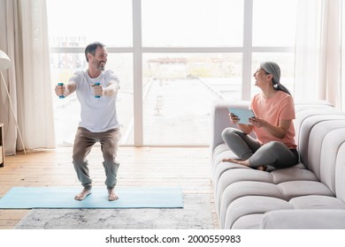 Mature husband doing sit-ups with dumbbells while his middle-aged wife watching fitness tutorial on tablet , avoiding physical exercises, being lazy at home on lockdown - Powered by Shutterstock