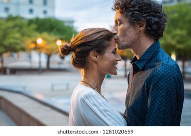 Mature husband kissing wife on forehead in the street in the evening. Romantic senior man giving a kiss to her woman in the city street. Loving middle aged couple in love at dusk.