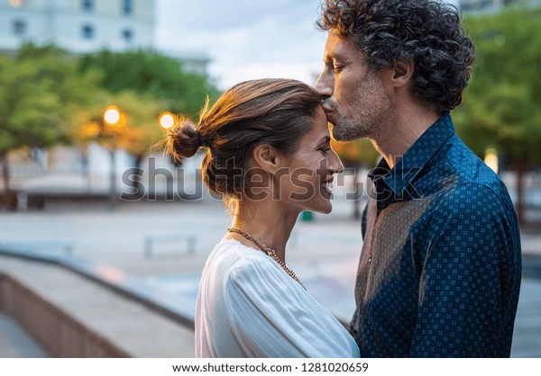 成熟的丈夫親吻妻子在街上的額頭在晚上 浪漫的高級男人給一個吻她的女人在城市街上 黃昏時相愛的中年夫婦 庫存照片