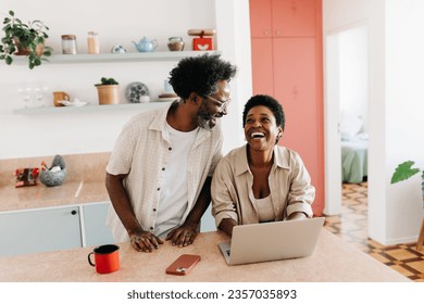 Mature husband and wife stand together in the kitchen, laughing while using a laptop. Happy black couple having fun together during their free time, enjoying technology and each other's company.