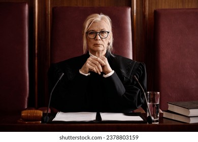 Mature impartial female judge in eyeglasses and black mantle looking at camera