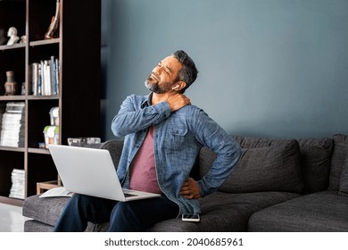 Mature indian man suffering from shoulder and back pain while sitting on couch and working from home on laptop. Stressed middle eastern businessman suffering from neck pain and stretching. - Shutterstock ID 2040685961