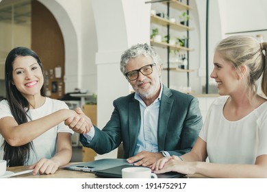 Mature Investor Shaking Hands With Young Entrepreneurs. Man And Women Meeting Over Cup Of Coffee At Co-working. Medium Shot. Partnership And Investment Concept