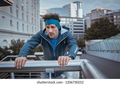 Mature Jogger With Headband Resting After Workout In A Winter Day. Tired Runner Taking Breath After Running In The City. Senior Sport Man Leaning Over Bridge Railing During Early Morning Workout.