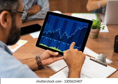 Mature latin businessman reading business graphs and stock charts on digital tablet. Rear view of business man holding tablet computer with stock analytics and abstract graphs. Close up hands working.