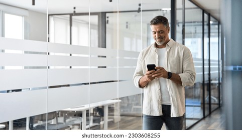 Mature Latin or Indian businessman walking with smartphone in office. Middle aged manager using cell phone mobile app. Digital technology application and solutions for business success development. - Powered by Shutterstock