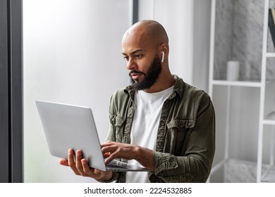 Mature Latin Male Freelancer Using Laptop Computer And Wearing Earbuds, Standing Near Window And Typing On Keyboard, Home Interior. Technology And Freelance Career Concept