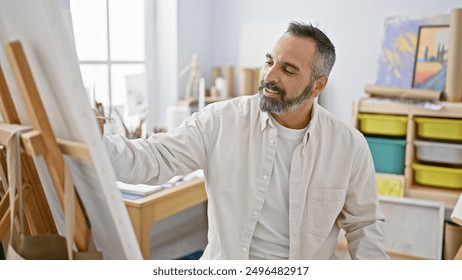 A mature latin man with grey beard smiles while painting in a well-lit art studio - Powered by Shutterstock