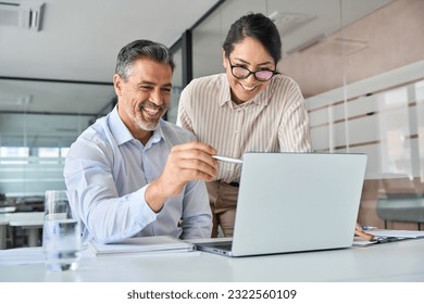 Mature Latin manager mentor talking to young Asian female coworker, showing online project results at meeting. Two happy diverse professional executives team working in office using pc laptop.